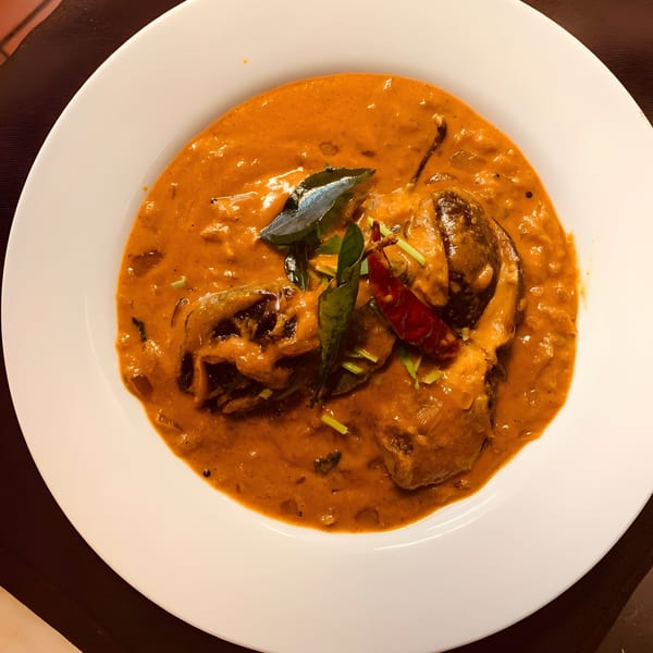 Aaha Indian Cuisine's eggplant curry, garnished with curry leaves and red chili, in a white bowl.